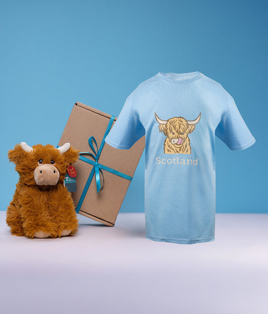Light blue t-shirt with a highland cow design and 'Scotland' text, next to a plush toy highland cow and gift box on a blue background.