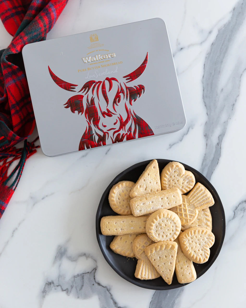 A tin of Walker's Highland Coo Icon Shortbread with a red-and-black plaid cow illustration, placed on a white marble surface next to a black plate of round and various shaped cookies and a red plaid scarf.