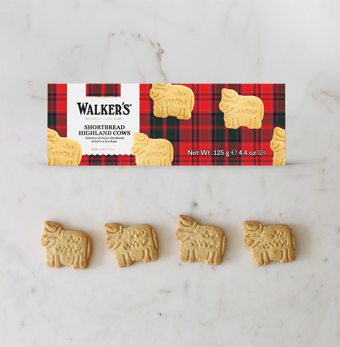 A package of Walker's Shortbread Highland Cows biscuits displayed on a white marble surface, with four small cow-shaped shortbread biscuits arranged below the box.