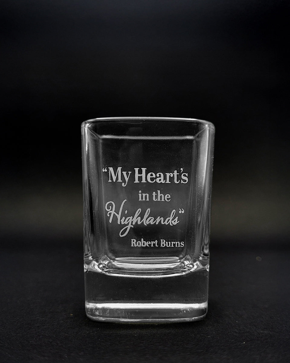 A clear glass shot glass with etched text reading “My Heart's in the Highlands” and “Robert Burns,” photographed against a dark black background.