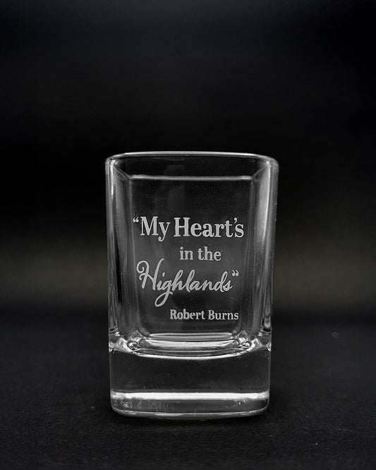 A clear glass shot glass with etched text reading “My Heart's in the Highlands” and “Robert Burns,” photographed against a dark black background.
