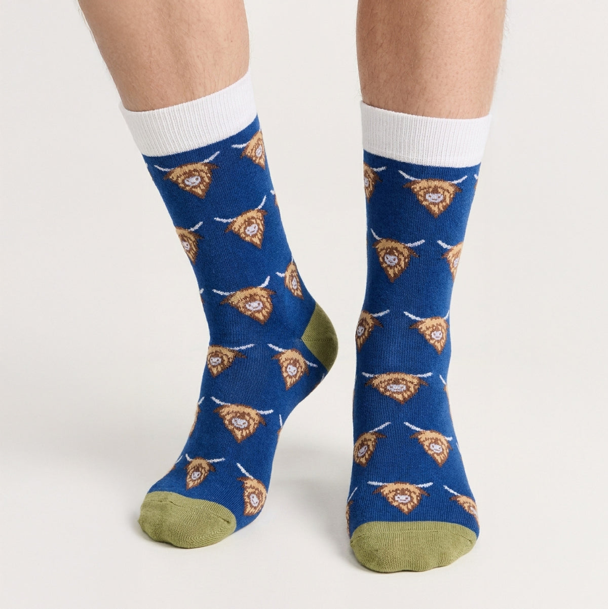 A pair of crew socks worn on a person's feet, featuring a blue background with a repeating pattern of tan and white highland cow heads, white ribbed cuffs, and olive green toes and heels.