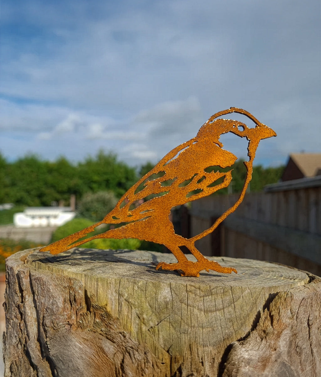 Rust-colored metal Sparrow fence topper sculpture on a wooden post with a blurred natural background