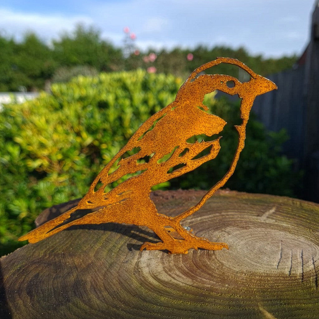 An orange rust-colored metal bird sculpture with cutout details, standing on a weathered wooden log with a blurred garden background.