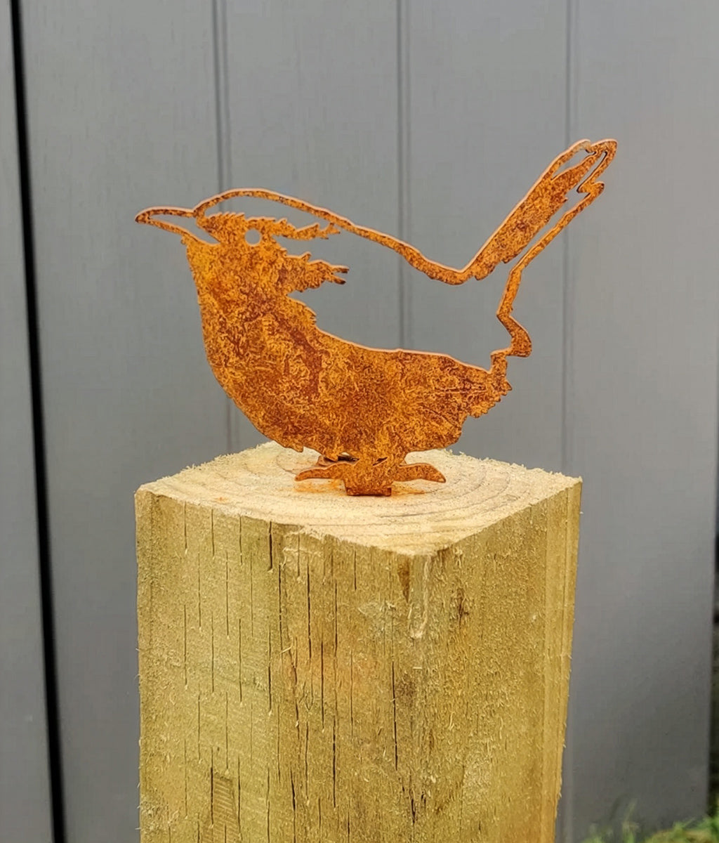 Rusted metal Wren Fence Topper sculpture on a wooden stump against a grey wall.