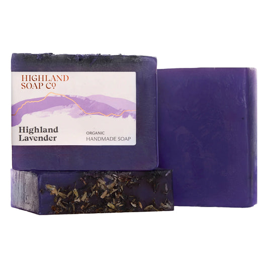 Three bars of Highland Soap Co. Highland Lavender organic handmade soap, showing the dark purple color, one with a paper label, and one with dried lavender buds embedded on the bottom, set against a plain white background.