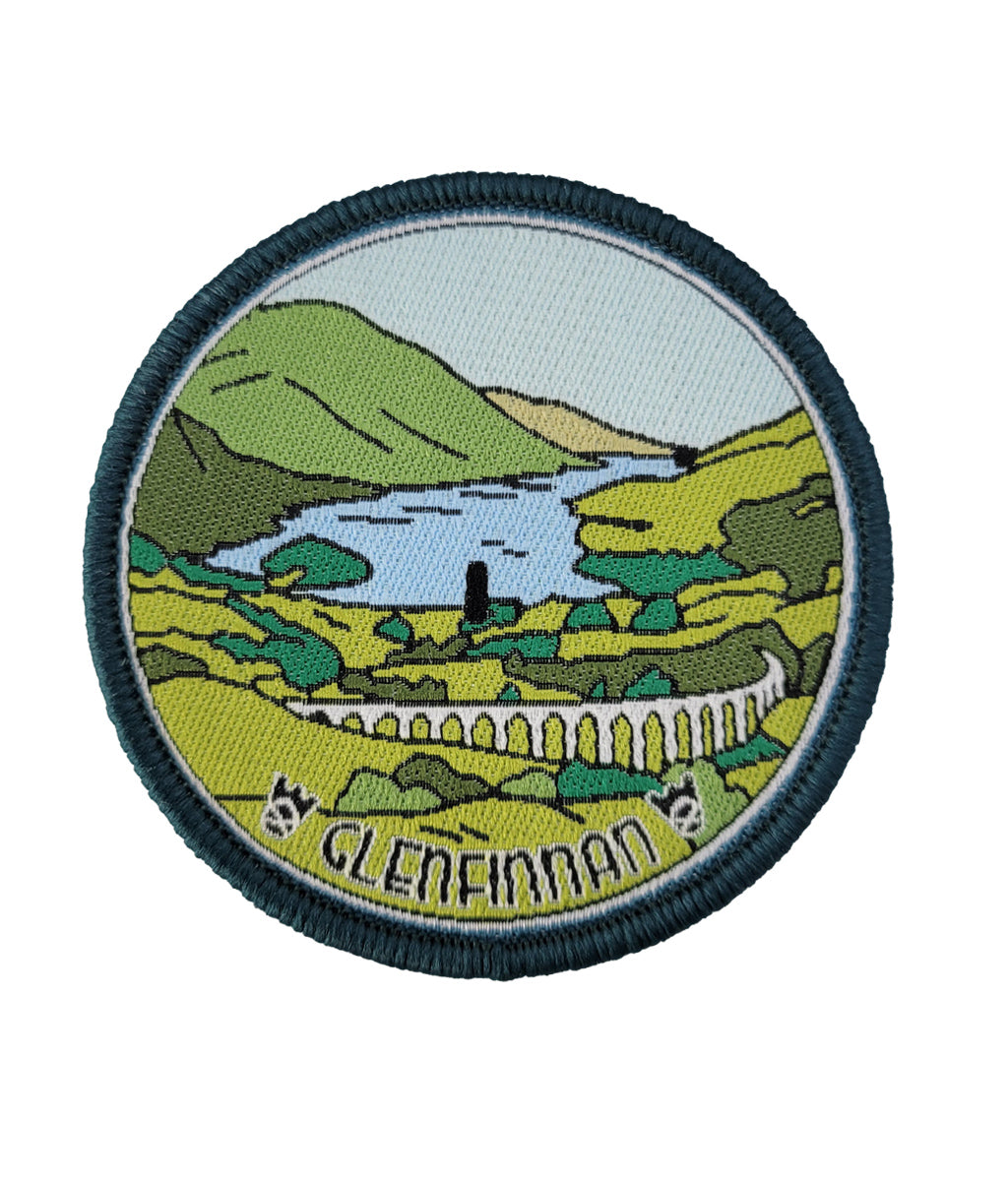 A circular embroidered patch depicting a scenic landscape labeled 'GLENFINNAN'. The patch features green hills, a light blue body of water, and the Glenfinnan Viaduct, all enclosed in a dark navy blue stitched border.