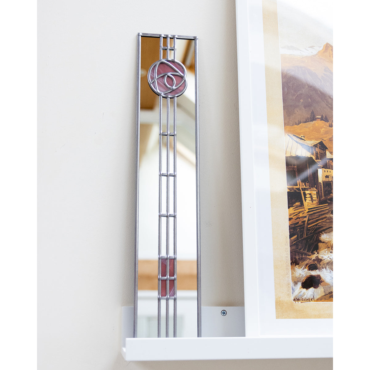 Tall, narrow mirror with decorative design on a white wall next to a framed picture.