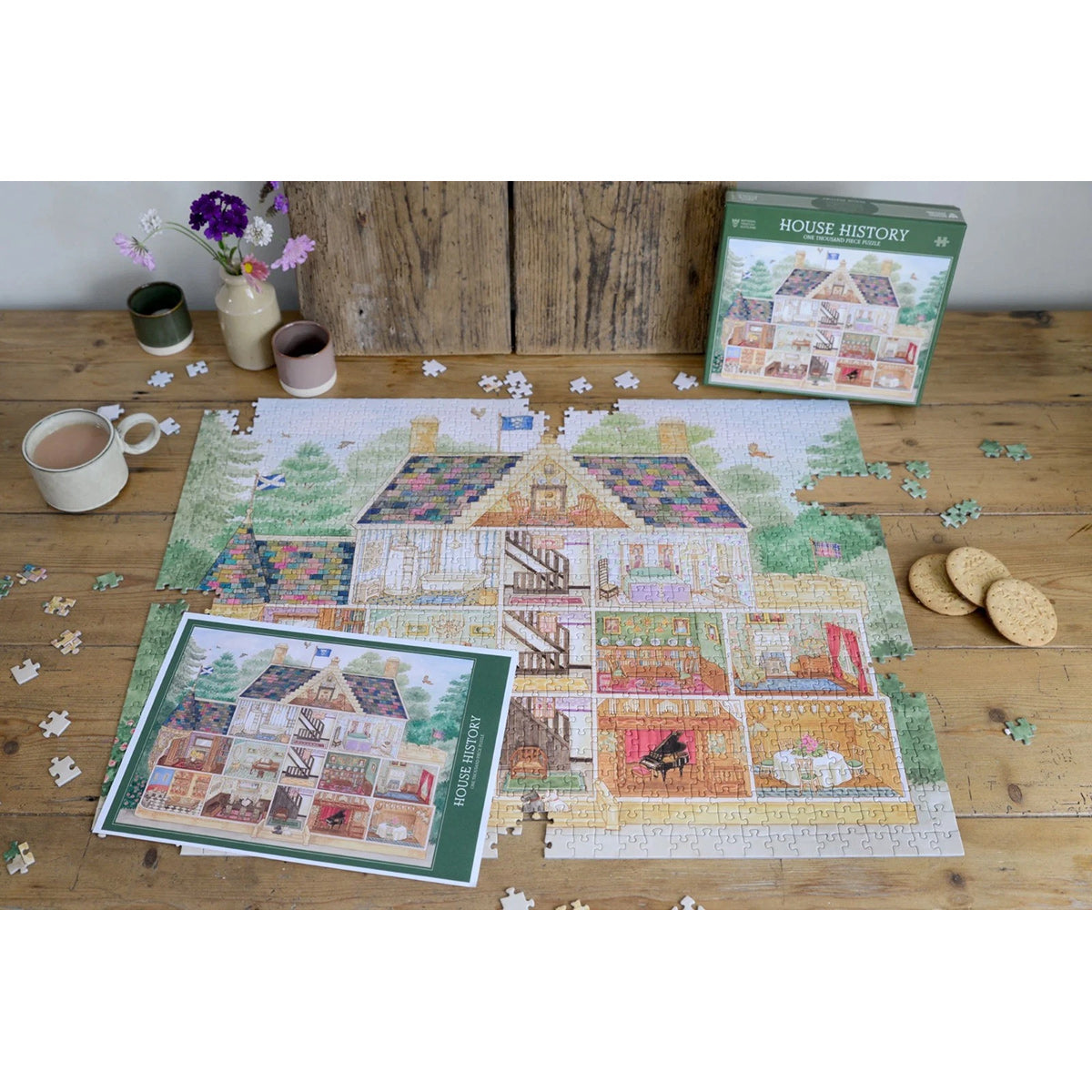 Jigsaw puzzle of a house with completed sections on a wooden table, surrounded by coffee, cookies, and flowers.