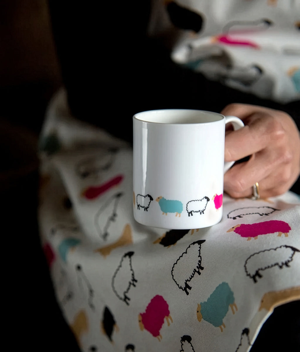 Person holding a white mug with colorful Woolly Ewe sheep designs on a patterned surface.