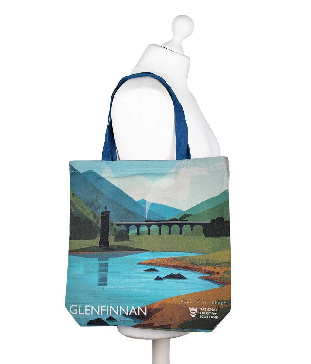 A canvas tote bag featuring a stylized landscape print of Glenfinnan landscape, Scotland, with blue handles and a scenic illustration of mountains, the Glenfinnan Viaduct, and monument.