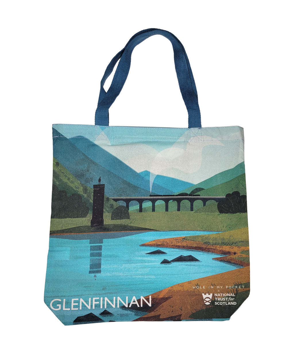 A canvas tote bag featuring a stylized landscape print of Glenfinnan landscape, Scotland, with blue handles and a scenic illustration of mountains, the Glenfinnan Viaduct, and monument.