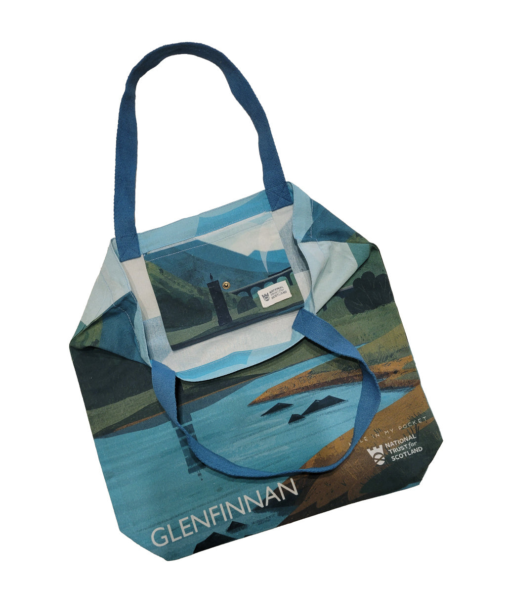 A canvas tote bag featuring a stylized landscape print of Glenfinnan landscape, Scotland, with blue handles and a scenic illustration of mountains, the Glenfinnan Viaduct, and monument.