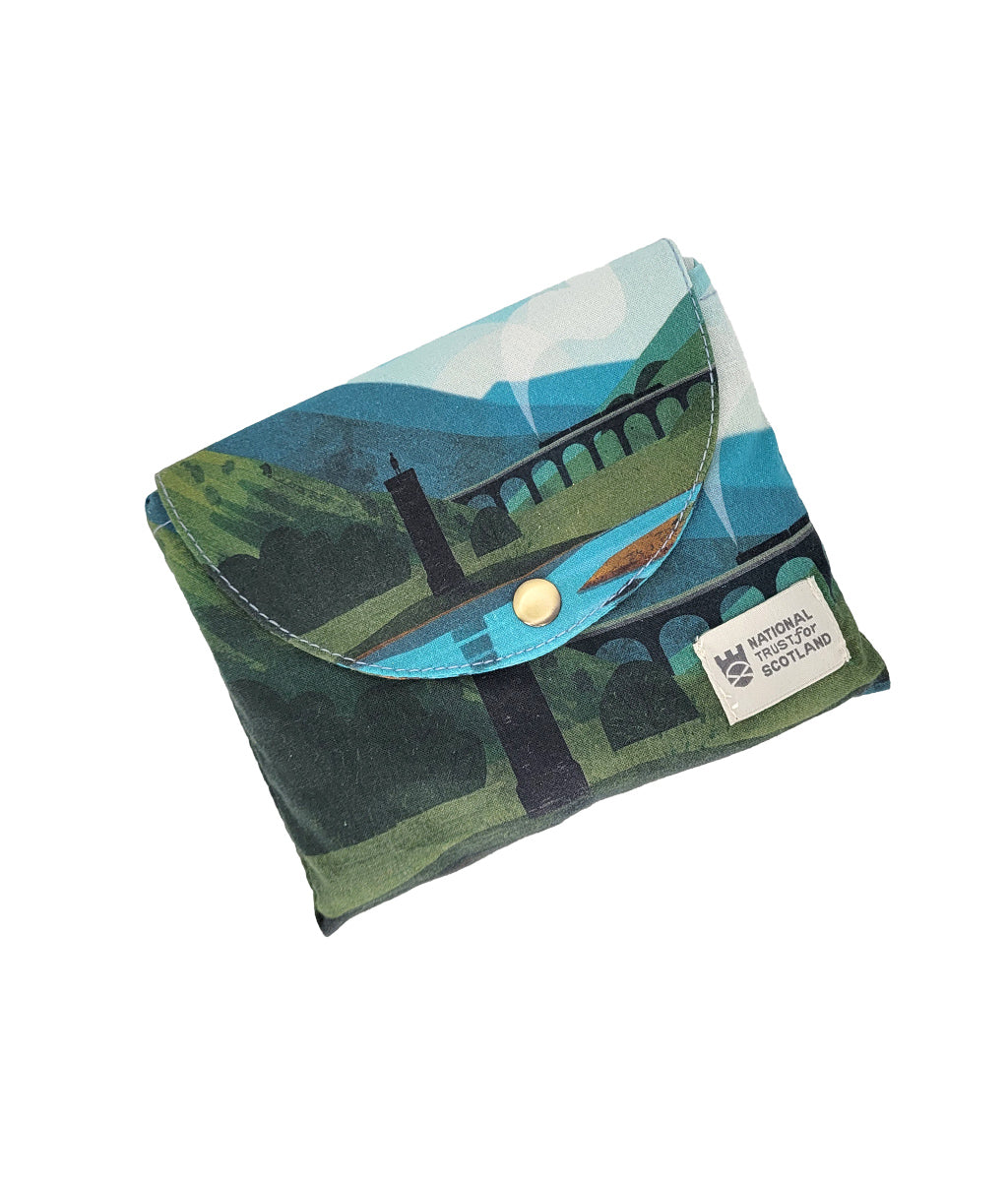 A small fabric pouch for the canvas tote bag featuring a stylized landscape print of Glenfinnan landscape, Scotland, with blue handles and a scenic illustration of mountains, the Glenfinnan Viaduct, and monument.