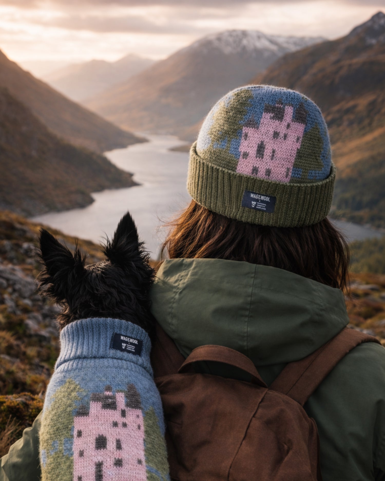 Person wearing a knit hat and mittens with a dog, both featuring castle designs, overlooking a mountainous landscape.