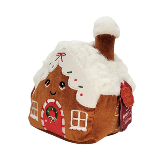 A gingerbread house plush toy with a smiling face, white faux-fur roof and chimney, a red door with a wreath, and a hanging red tag.
