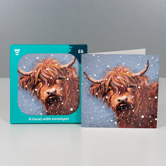 A set of illustrated greeting cards with matching envelopes, shown with the turquoise card box and one card standing open. The artwork features a shaggy Highland cow in warm brown tones against a snowy blue background.