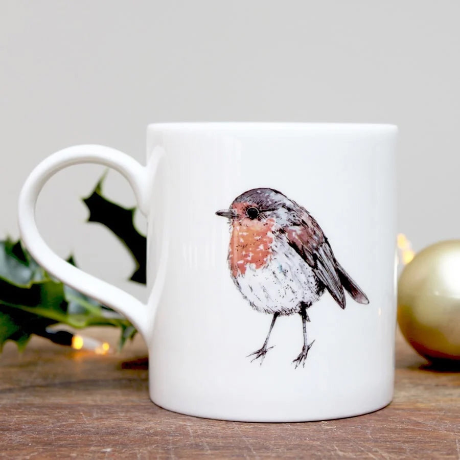 A white ceramic mug featuring a detailed illustration of a robin bird with a rust-red breast, black head, and brown wings. The mug is set on a wooden surface with a blurred background containing holly leaves, warm string lights, and a gold ornament.