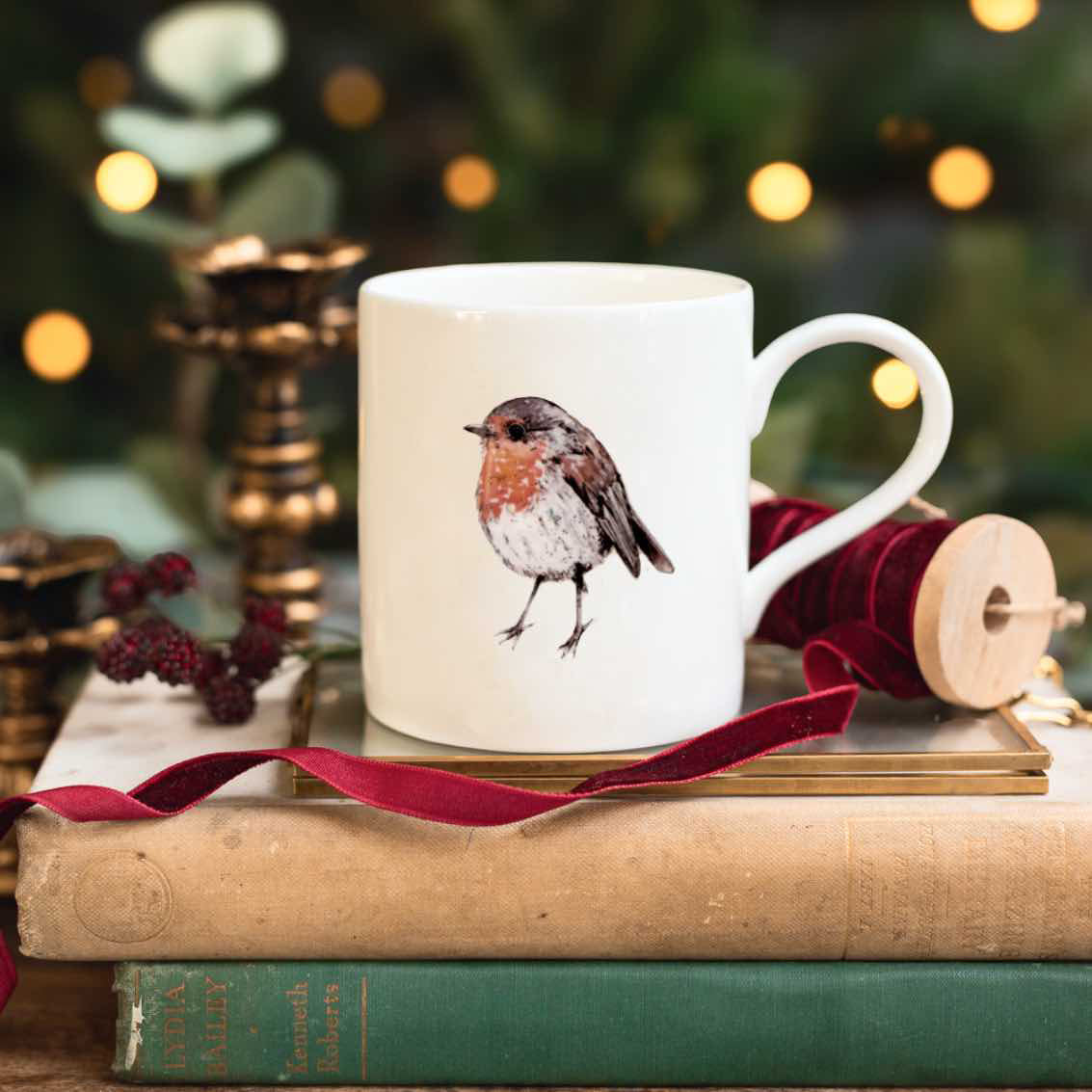 A white ceramic mug with a watercolor robin bird illustration, styled on a stack of vintage books with a red ribbon and holiday decorations in the background.