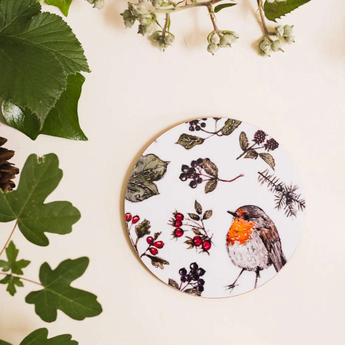 A round wooden coaster with a white background printed with a robin bird and winter botanical illustrations, styled on a cream surface with green leaves and small white flowers.