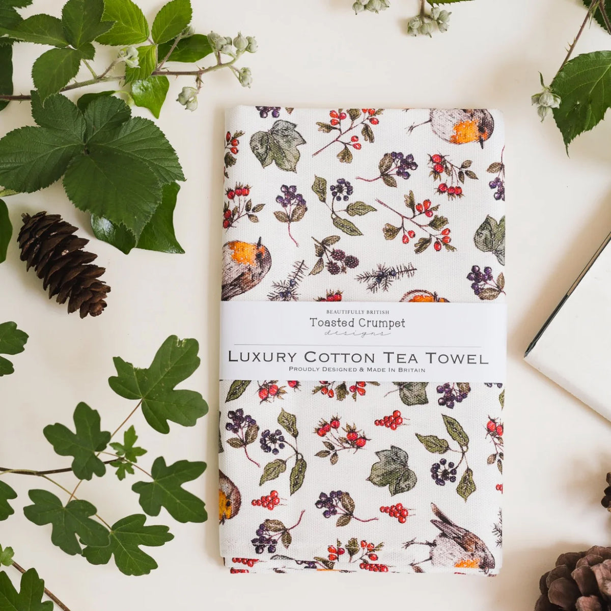 A folded cotton tea towel with a floral and Robin print, wrapped in a white paper band labeled 'Luxury Cotton Tea Towel' and 'Toasted Crumpet Designs,' styled on a light surface with green leaves, a pinecone, and part of a book.