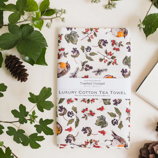 A folded cotton tea towel with a floral and Robin print, wrapped in a white paper band labeled 'Luxury Cotton Tea Towel' and 'Toasted Crumpet Designs,' styled on a light surface with green leaves, a pinecone, and part of a book.