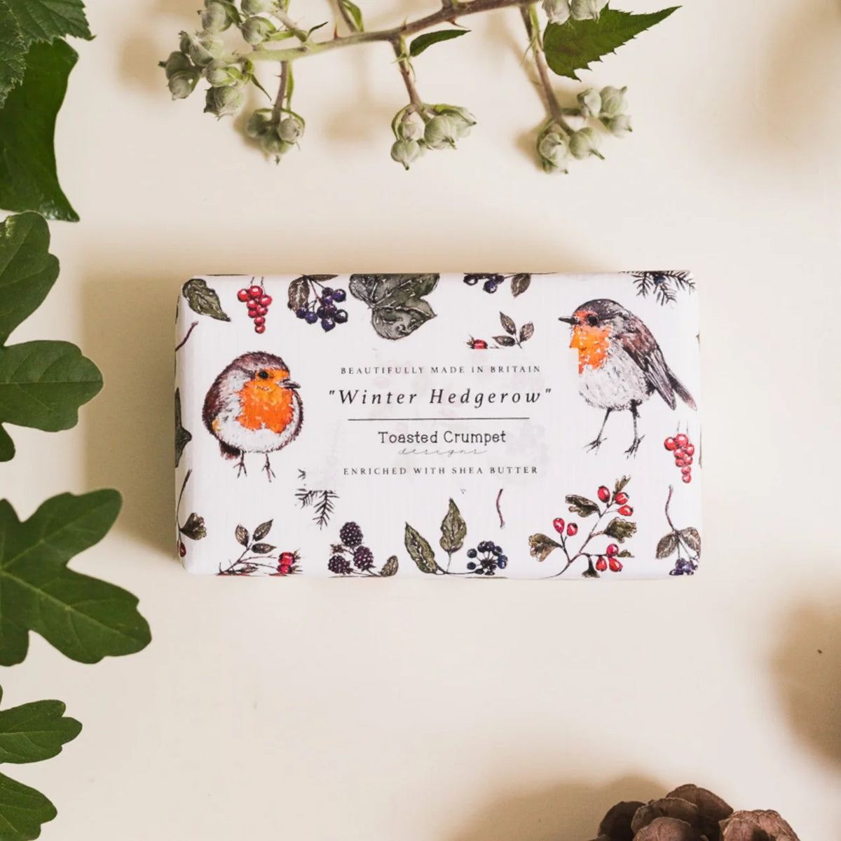 A rectangular bar of soap wrapped in white paper printed with robin birds and winter berries, photographed on a warm cream background with decorative leaves and small flowers around it.