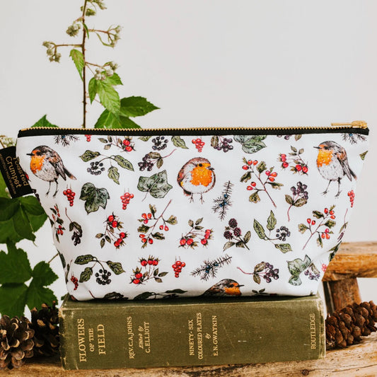 A white zip pouch with a detailed robin and holly print, styled on top of a vintage green book with pinecones and greenery in the background.