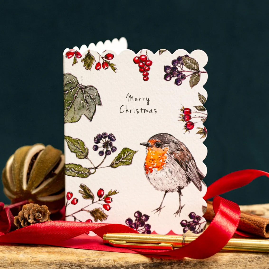A scalloped-edge Christmas greeting card featuring a robin bird and winter berries, styled on a wooden surface with a red ribbon, gold pen, and pinecones against a dark teal background.
