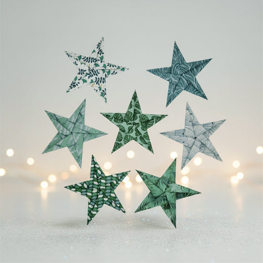 Six decorative paper star ornaments in assorted green, teal, gray, and white leaf-patterned designs, arranged against a soft light gray background with blurred warm string lights.
