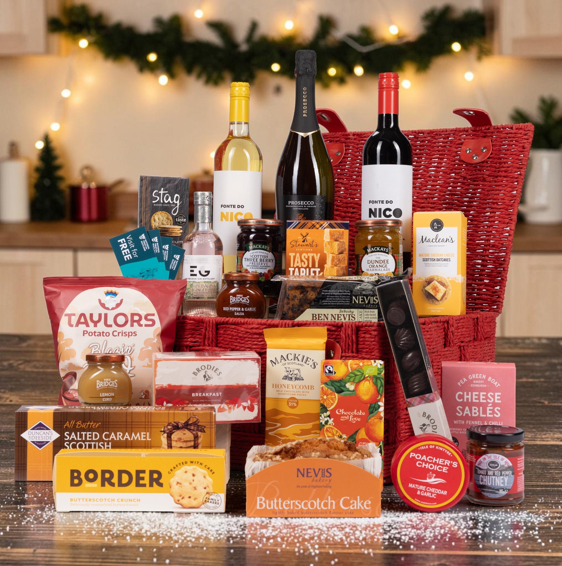 A Christmas-themed gourmet gift hamper filled with an assortment of British snacks, cheeses, and beverages, arranged in a red woven basket on a wooden table with festive lights in the background.