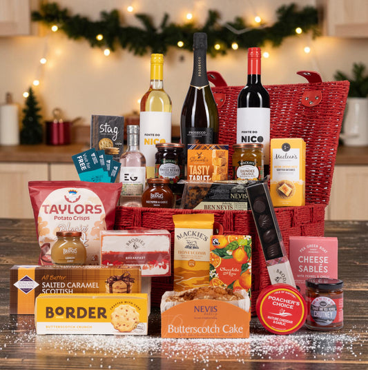 A Christmas-themed gourmet gift hamper filled with an assortment of British snacks, cheeses, and beverages, arranged in a red woven basket on a wooden table with festive lights in the background.