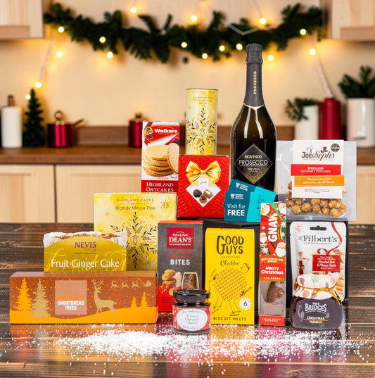 A Christmas hamper gift basket filled with assorted gourmet treats, snacks, and a bottle of Prosecco, arranged on a wooden table with a festive kitchen background decorated with lights and greenery.