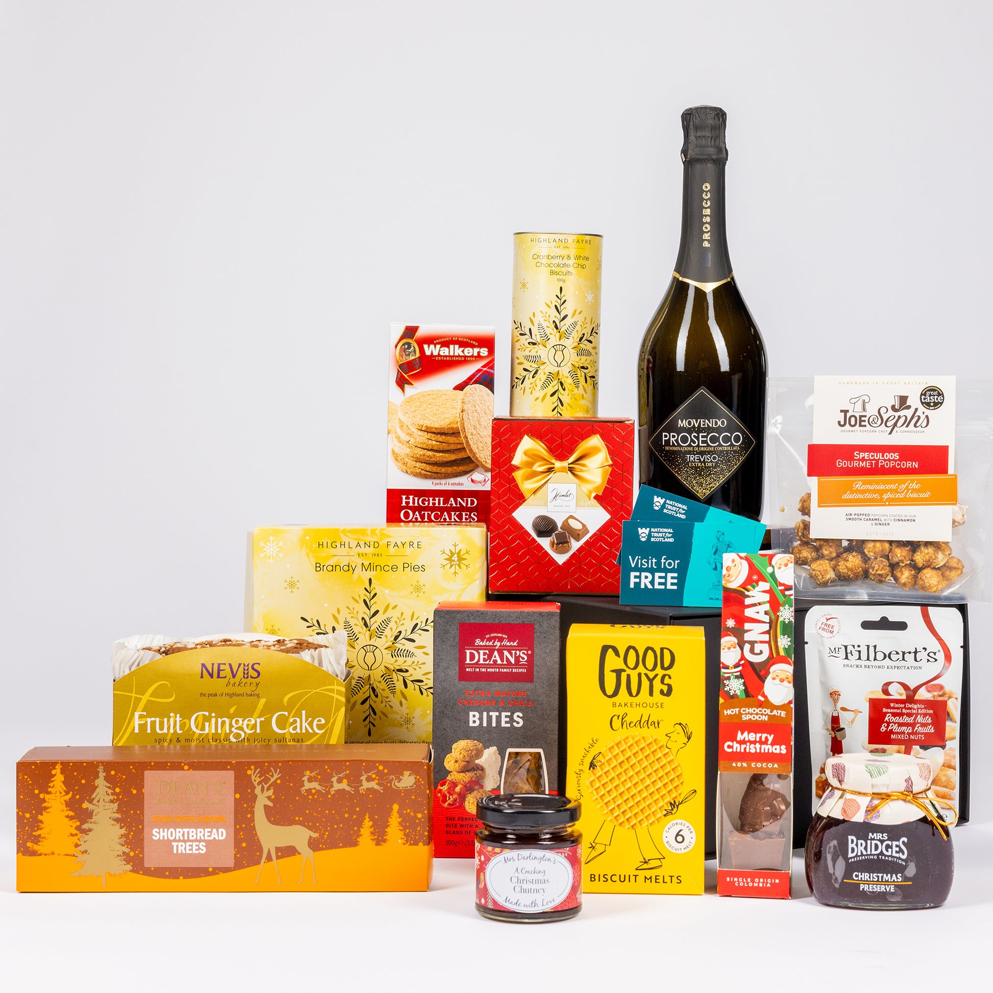 A curated gift hamper featuring an assortment of food products, snacks, and a bottle of Prosecco arranged together on a light gray background.
