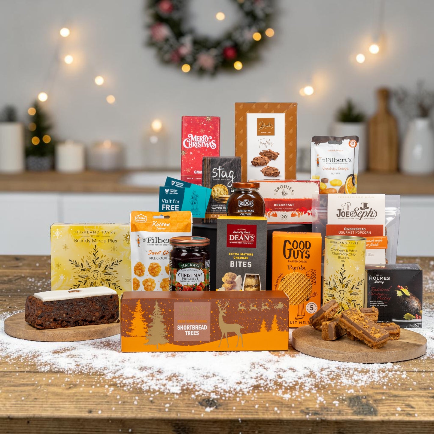 A Christmas gift hamper filled with assorted UK snacks, sweets, and tea arranged on a wooden table with a white powder snow dusting, set against a softly blurred festive background with candles and a wreath.