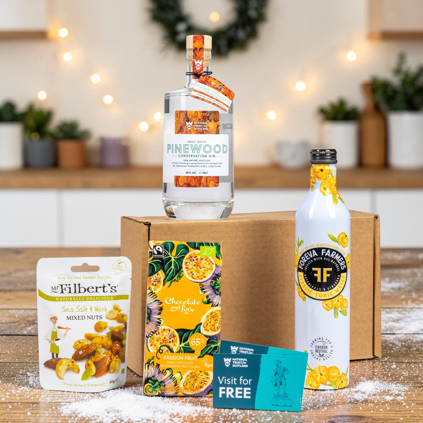 A curated gift set arranged on a wooden table in front of a cardboard box, featuring a clear bottle of Pinewood Gin with a warm blurred kitchen background.