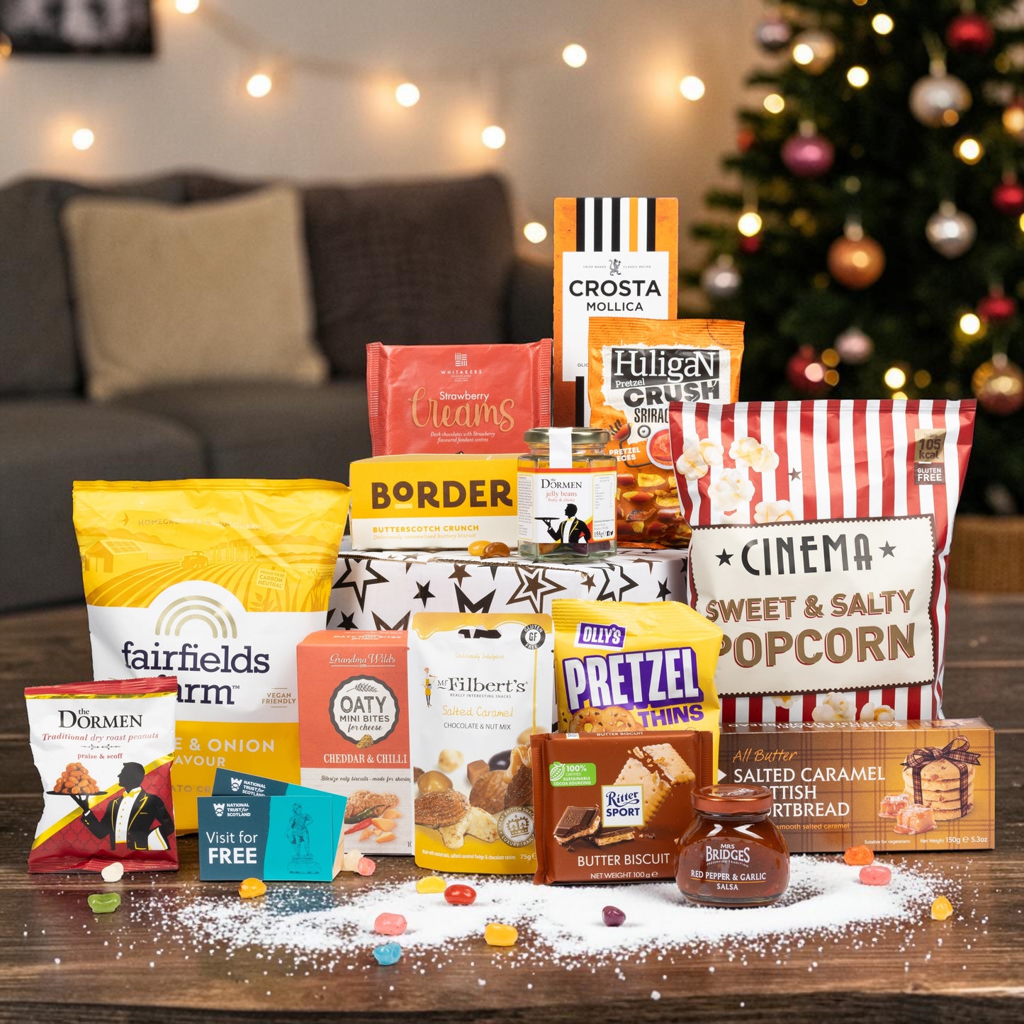 A movie night-themed gourmet snack and candy gift basket arranged on a wooden table in front of a blurred living room background with a lit Christmas tree. 