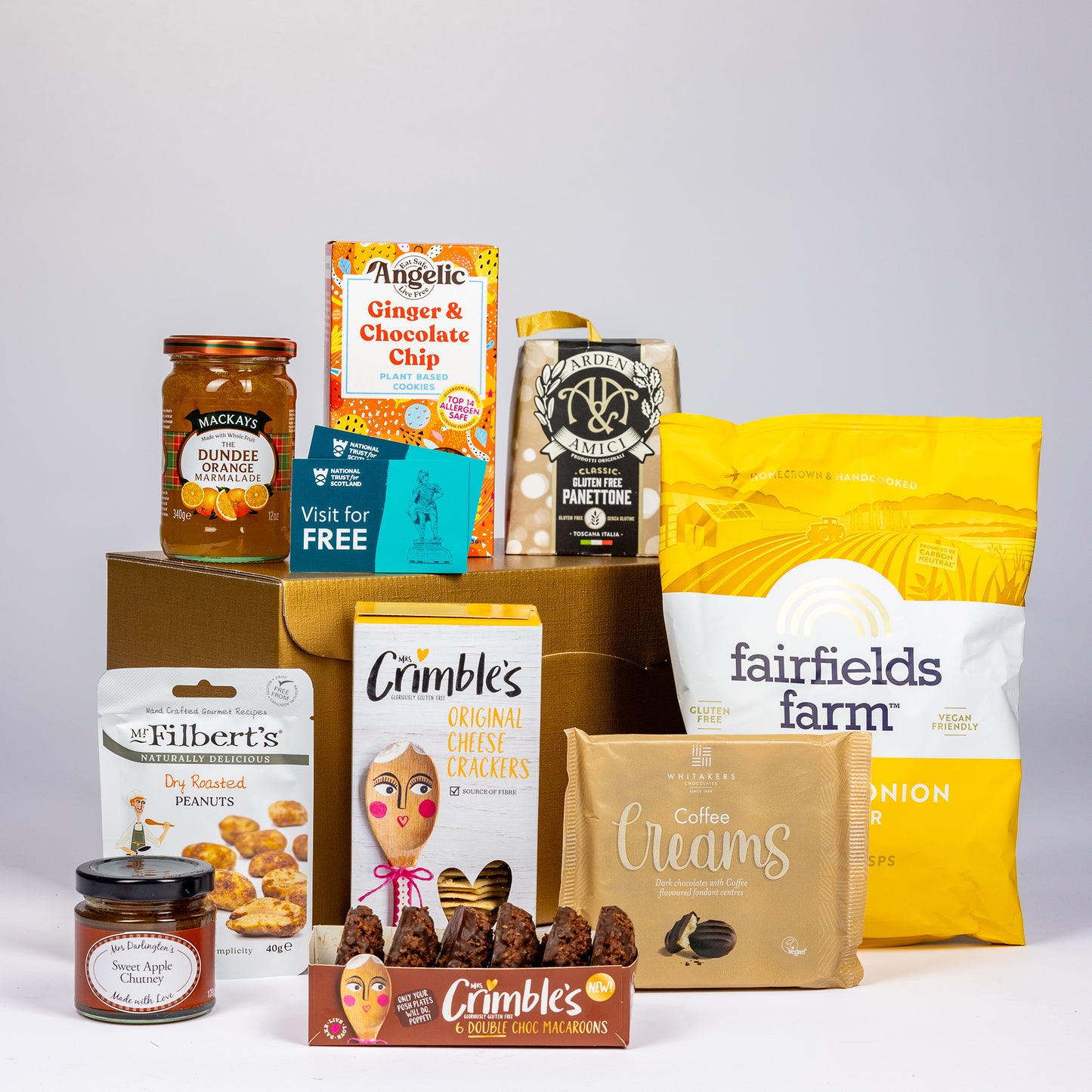 A curated gift basket assortment of snacks and treats arranged on a white surface with a gold cardboard box behind them. 