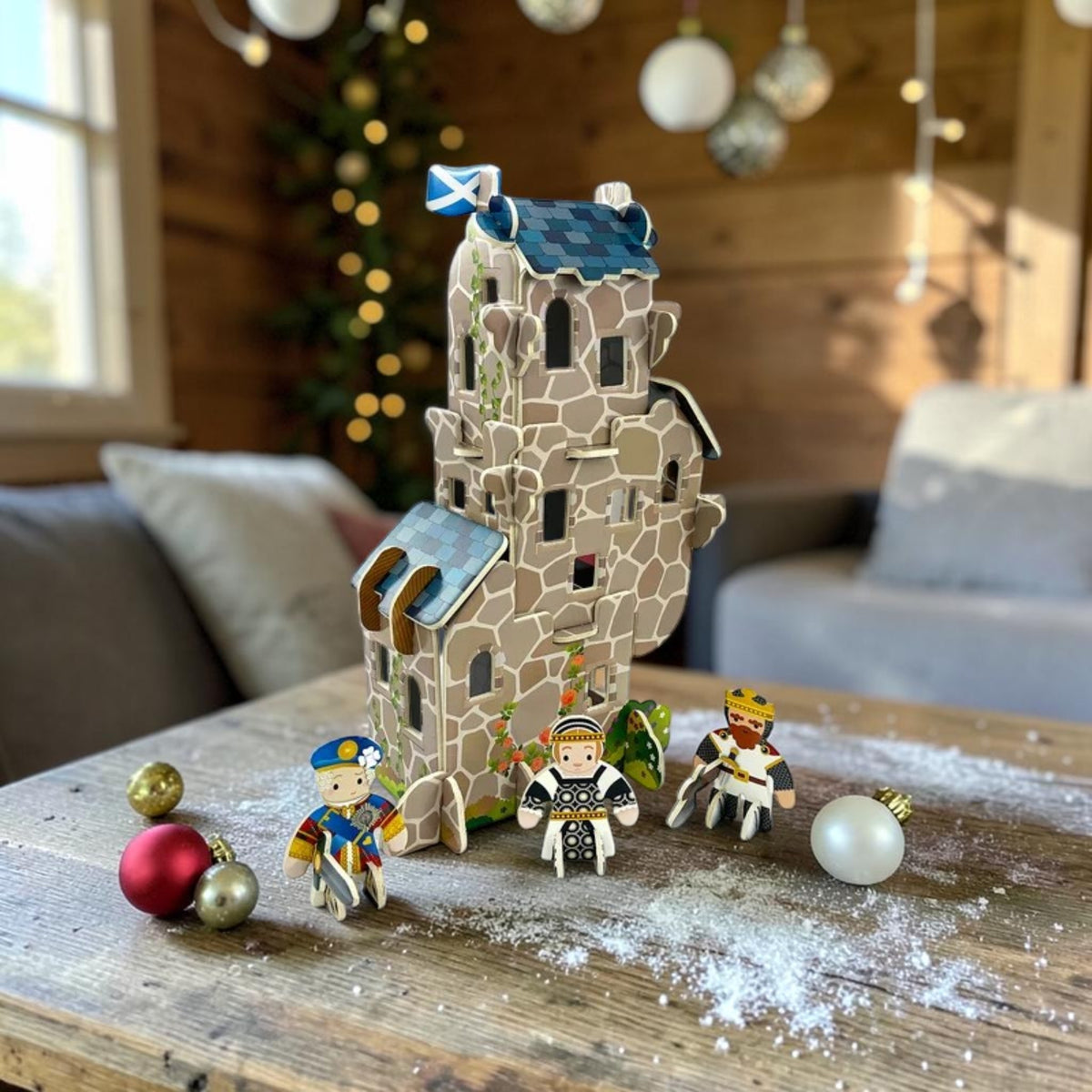 A decorative miniature castle model with a Scottish flag on top, displayed on a wooden table in a cozy living room setting with small toy figures and Christmas ornaments.
