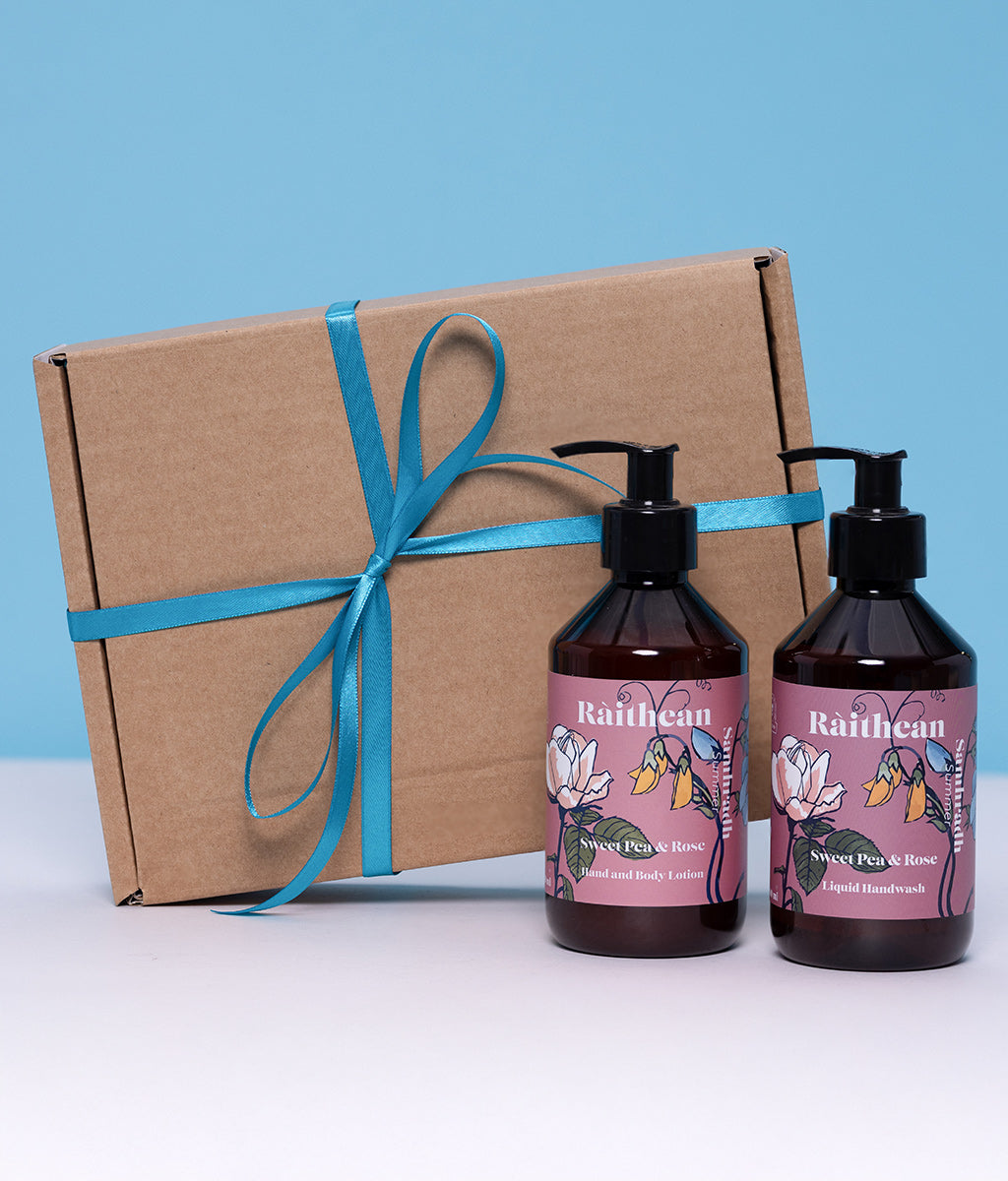 Two bottles of Raithean Sweet Pea & Rose hand and body wash with a brown box tied with teal ribbon on a light blue background.