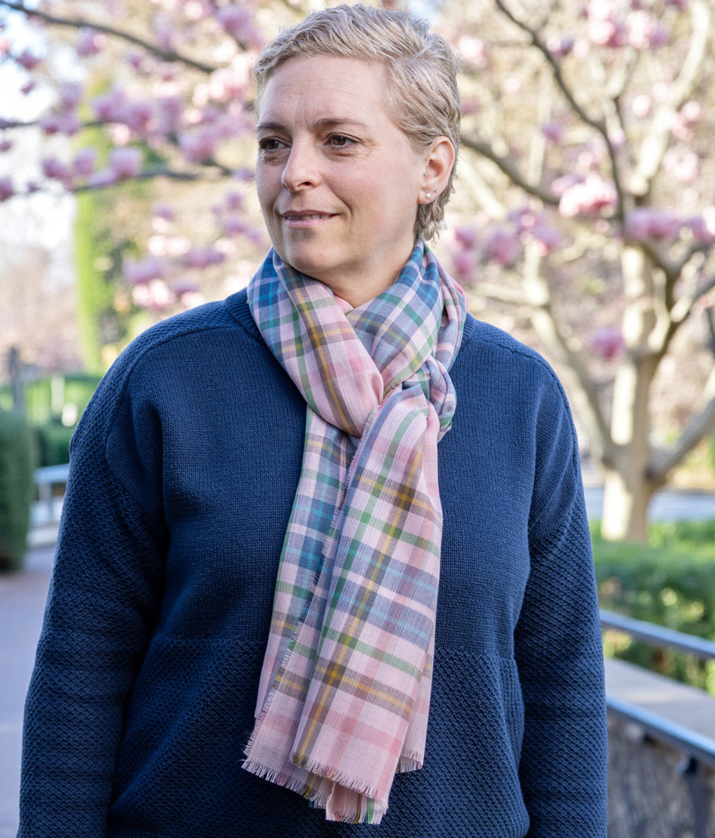 Person wearing a blue sweater and a Pink Castle Tartan Merino Wool Scarf with cherry blossom trees in the background