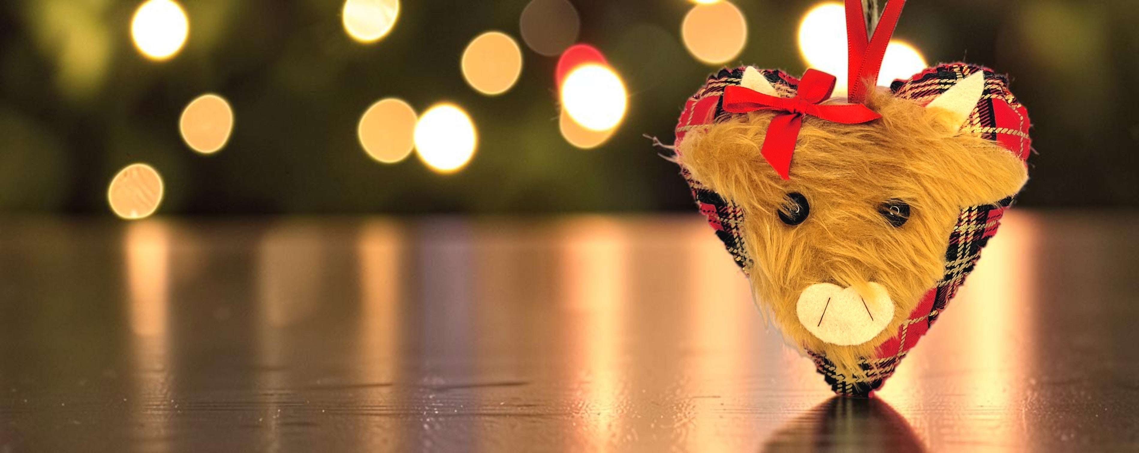 A heart-shaped Christmas ornament inspired by a highland cow, made of tan faux fur and wrapped in a red plaid fabric, with a red ribbon bow and hanging loop, set against a dark background with warm bokeh lights.