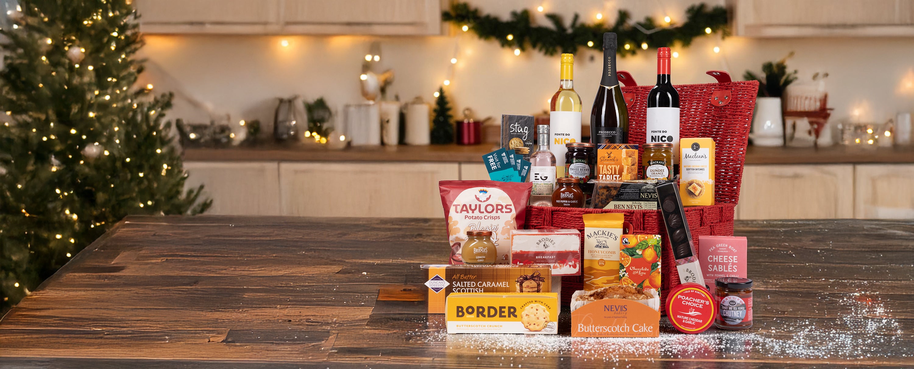 A Christmas-themed gourmet gift basket arranged on a wooden table in a warmly lit kitchen. The basket contains assorted packaged foods, crackers, cheeses, and bottles, with a red patterned gift bag and a red basket base.