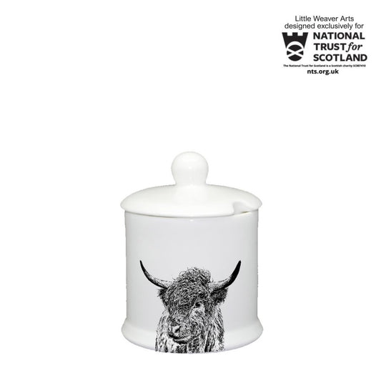 A white ceramic jar with a matching lid and a black-and-white highland cow illustration on the front, shown on a white background. A National Trust for Scotland badge appears in the top right corner.