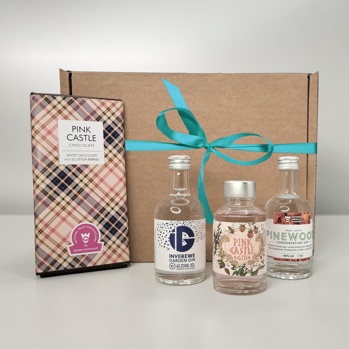 Three miniature gin bottles in a cardboard gift box with a teal ribbon, accompanied by a chocolate box labeled 'Pink Castle'.