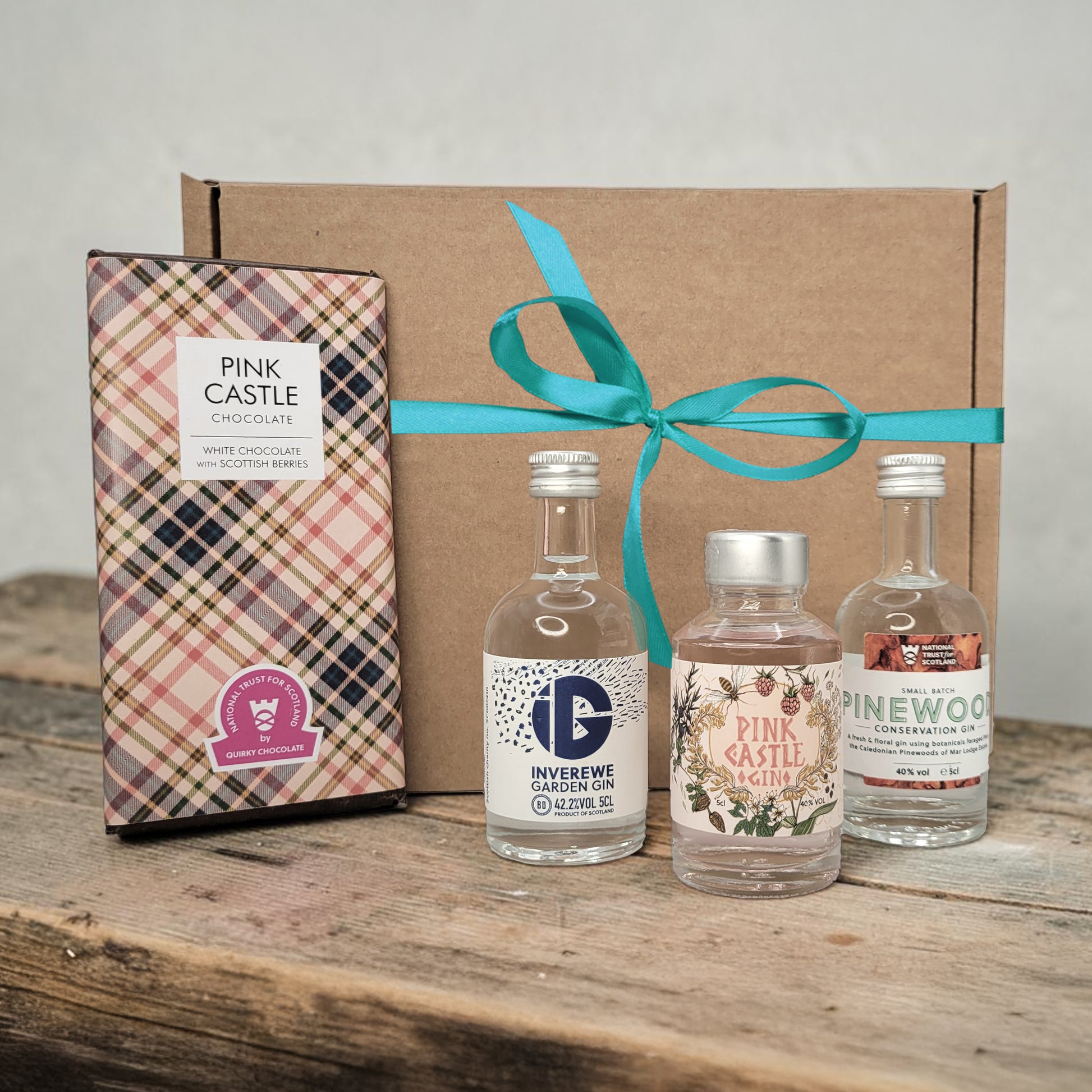 Three miniature gin bottles in a cardboard gift box with a teal ribbon, accompanied by a chocolate box labeled 'Pink Castle'.