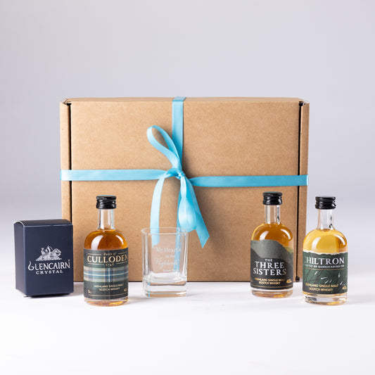 A whisky gift set featuring three mini single malt bottles and a shot glass from Glencairn Crystal arranged in front of a brown cardboard gift box tied with a teal ribbon on a light gray background.