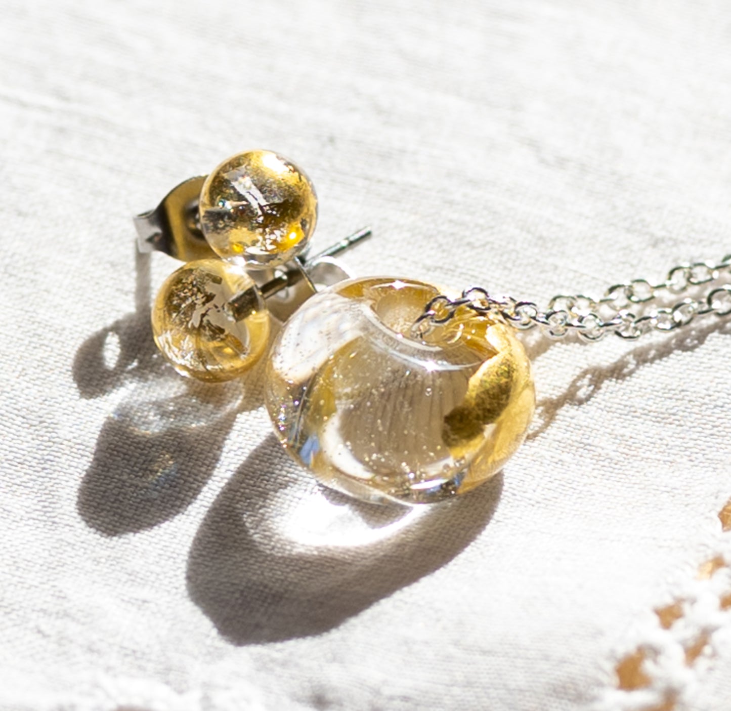 Recycled whisky bottle clear glass and gold pendant and matching clear glass stud earrings on a silver chain, displayed on a textured white fabric under bright sunlight.