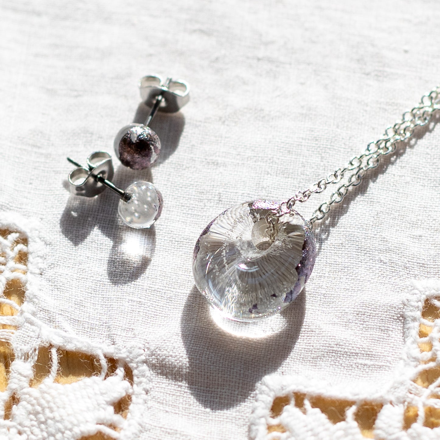 A recycled whisky bottle clear glass and palladium pendant and matching stud earrings, displayed on a textured white fabric with a lace border.