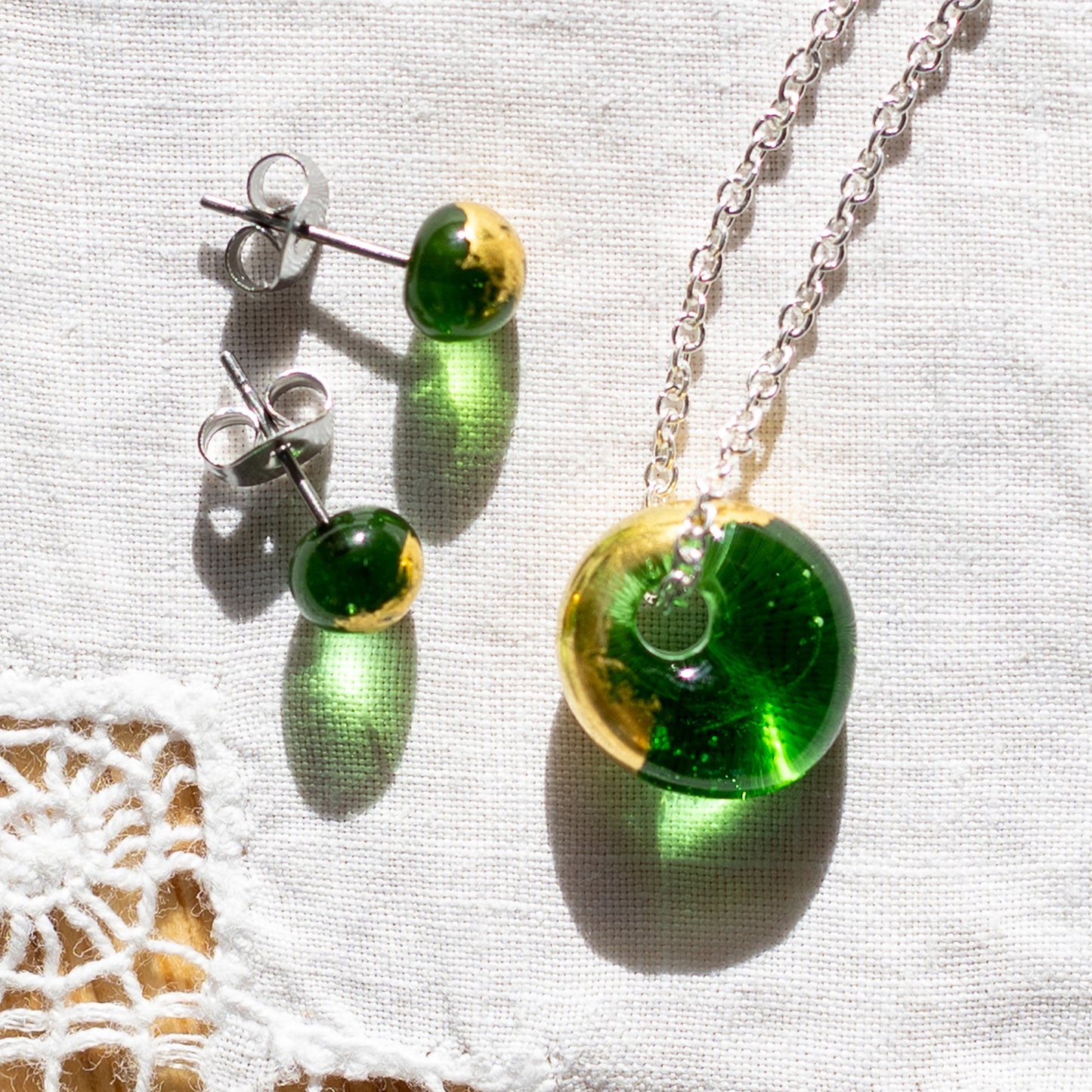 A jewellery set featuring a silver chain necklace and matching stud earrings with round green and gold glass pendant, displayed on a textured white fabric background.